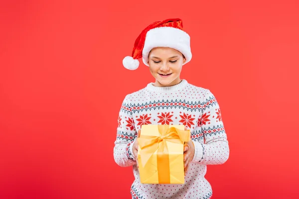 Front view of smiling kid in santa hat holding present isolated on red — Stock Photo