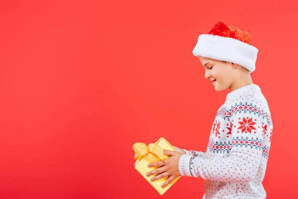 Вид збоку усміхненої дитини в капелюсі Санта тримає подарунок ізольовано на червоному — стокове фото