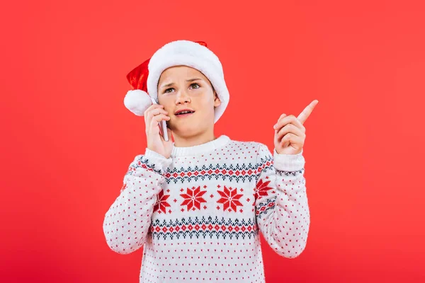 Niño disgustado en sombrero de santa hablar en el teléfono inteligente aislado en rojo - foto de stock