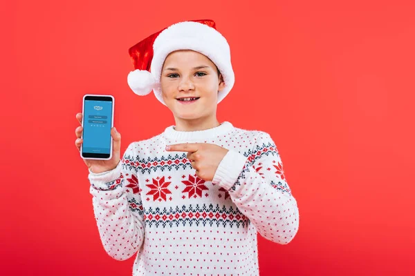 KYIV, UCRANIA - 9 DE SEPTIEMBRE DE 2019: vista frontal del niño sonriente en sombrero de santa señalando con el dedo al teléfono inteligente con la aplicación skype en la pantalla - foto de stock