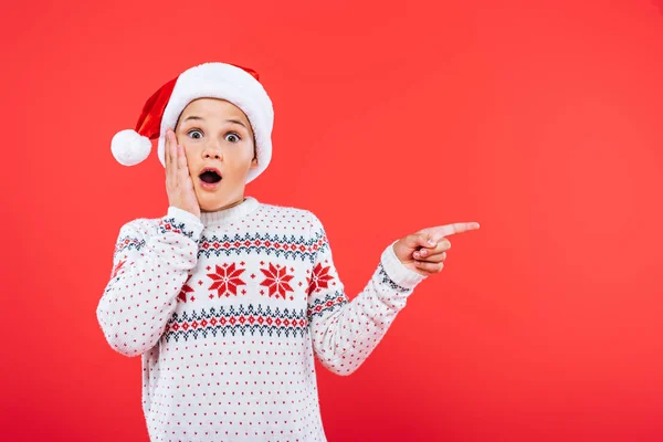 Front view of surprised kid in santa hat pointing with finger isolated on red — Stock Photo