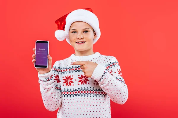 KYIV, UCRANIA - 9 DE SEPTIEMBRE DE 2019: vista frontal del niño sonriente en sombrero de santa señalando con el dedo al teléfono inteligente con la aplicación instagram en la pantalla - foto de stock