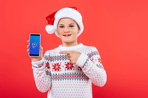 KYIV, UKRAINE - SEPTEMBER 9, 2019: front view of smiling kid in santa hat pointing with finger at smartphone with shazam app on screen — Stock Photo