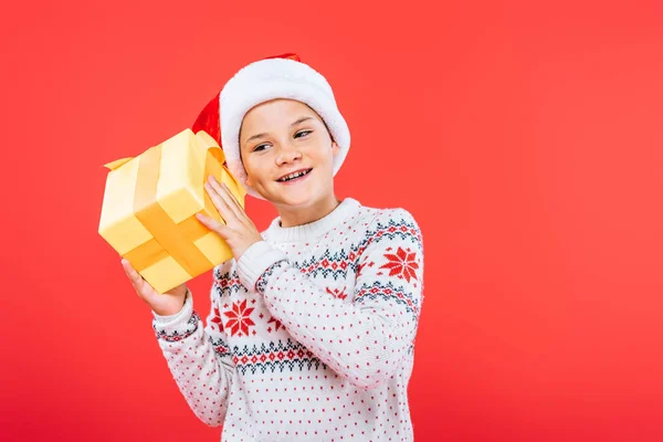 Niño sonriente en sombrero de santa celebración presente aislado en rojo - foto de stock