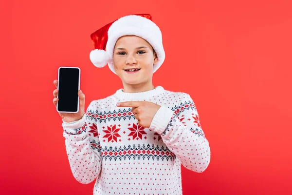 Vue de face de l'enfant souriant en santa chapeau pointant avec le doigt au smartphone avec écran blanc isolé sur rouge — Photo de stock
