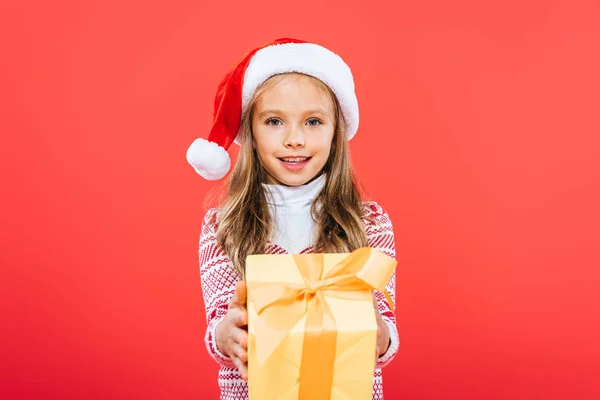 Front view of smiling kid in santa hat holding present isolated on red — Stock Photo