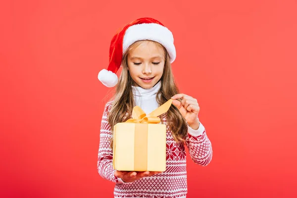 Усміхнена дитина в капелюсі Санта тримає подарунок ізольовано на червоному — стокове фото
