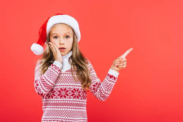 Front view of surprised kid in santa hat pointing with finger isolated on red — Stock Photo