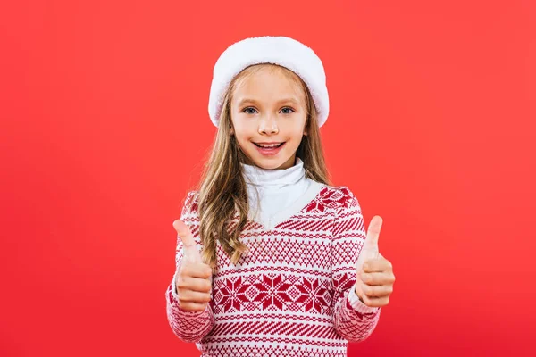 Vista frontal de niño sonriente en suéter y sombrero de santa que muestra los pulgares hacia arriba aislado en rojo - foto de stock