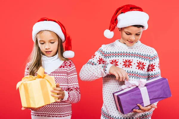 Vista frontal de dos niños sonrientes en sombreros de santa con regalos aislados en rojo - foto de stock