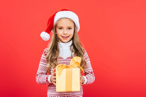 Front view of smiling kid in santa hat holding present isolated on red — Stock Photo