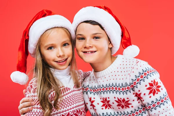 Front view of two smiling kids in santa hats and sweaters embracing isolated on red — стоковое фото