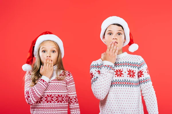 Front view of two shocked kids in santa hats and sweaters covering mouths with hands isolated on red — Stock Photo