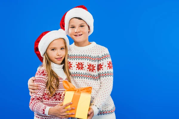 Two smiling kids in santa hats with present embracing isolated on blue — Stock Photo