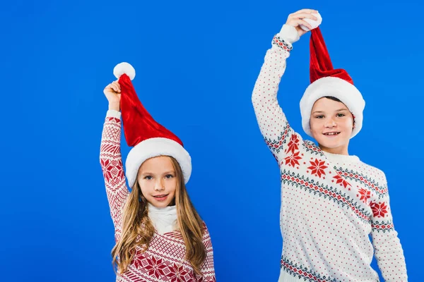 Вид спереди двух улыбающихся детей в свитерах и санта-хатах, изолированных на голубом фоне — стоковое фото