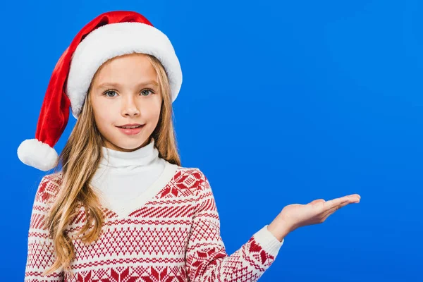 Frontansicht eines Kindes mit Weihnachtsmütze, das mit einer Hand isoliert auf blau zeigt — Stockfoto