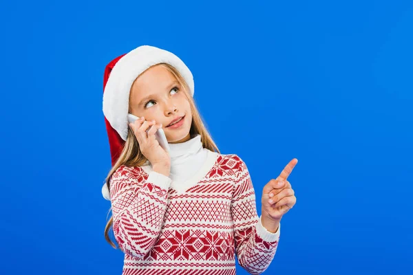 Kid in santa hat and sweater talking on smartphone isolated on blue — Stock Photo