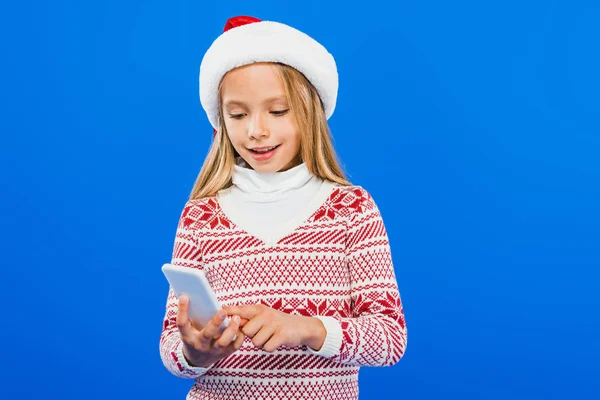 Smiling kid in santa hat and sweater using smartphone isolated on blue — Stock Photo