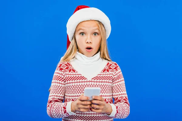 Vue de face de l'enfant choqué dans le chapeau santa et le pull en utilisant un smartphone isolé sur bleu — Photo de stock
