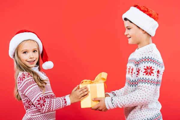 Dos niños sonrientes en sombreros de santa con presente aislado en rojo - foto de stock