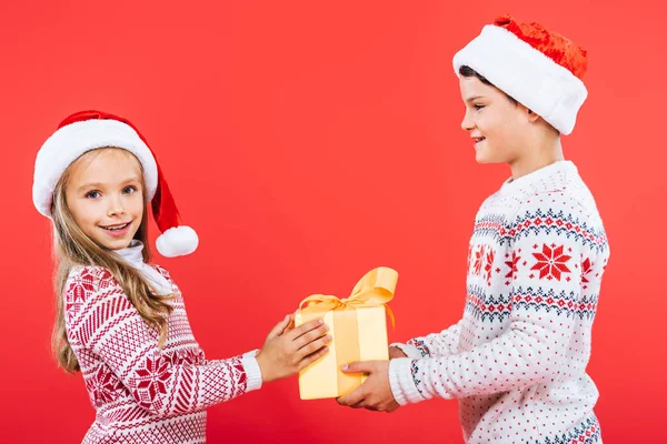 Dos niños sonrientes en sombreros de santa con presente aislado en rojo — Stock Photo