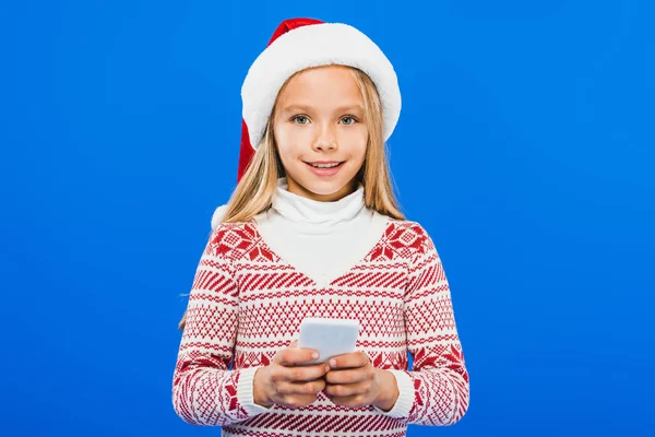 Front view of kid in santa hat and sweater using smartphone isolated on blue — Stock Photo