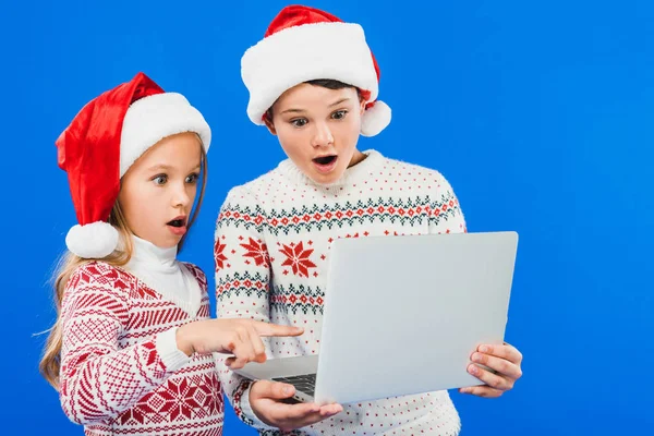 Dos niños sorprendidos en sombreros de santa usando el ordenador portátil aislado en azul - foto de stock