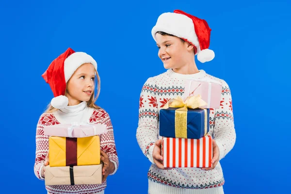 Due bambini sorridenti in cappelli da Babbo Natale con regali che si guardano isolati sul blu — Foto stock