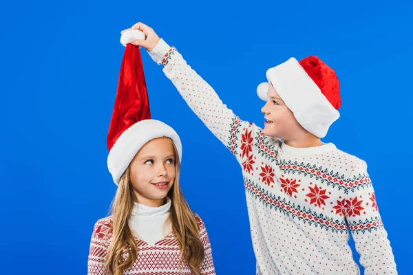 Zwei lächelnde Kinder in Weihnachtsmützen und Pullovern isoliert auf blauem Grund — Stockfoto