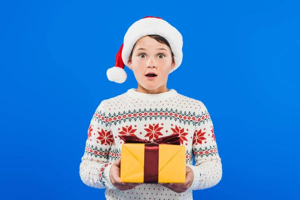 Vue de face de l'enfant choqué dans le chapeau de Père Noël et pull tenant cadeau isolé sur bleu — Photo de stock