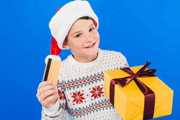 Smiling kid in santa hat holding gift and credit card isolated on blue — Stock Photo