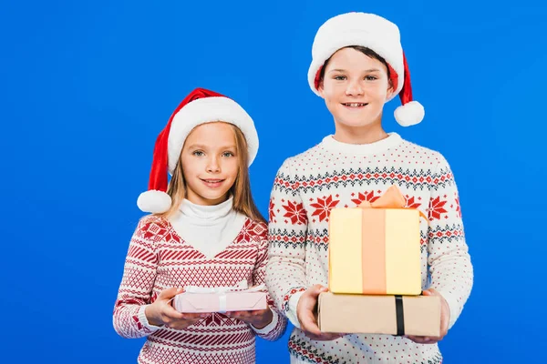 Vista frontal de dos niños sonrientes en sombreros de santa con regalos aislados en azul - foto de stock