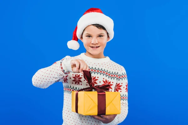 Vista frontale del bambino in cappello di Babbo Natale e maglione in possesso di regalo isolato su blu — Foto stock