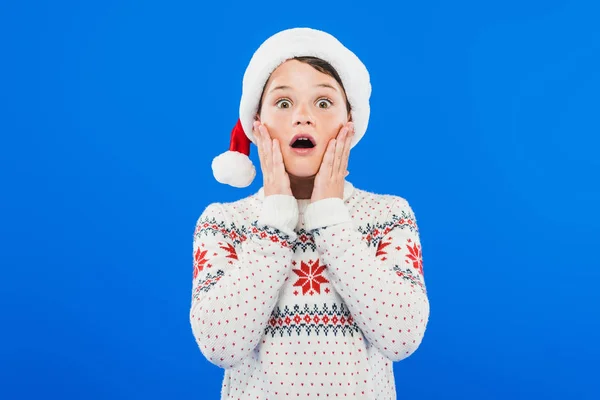 Vista frontale del bambino scioccato in cappello di Babbo Natale e maglione isolato su blu — Foto stock