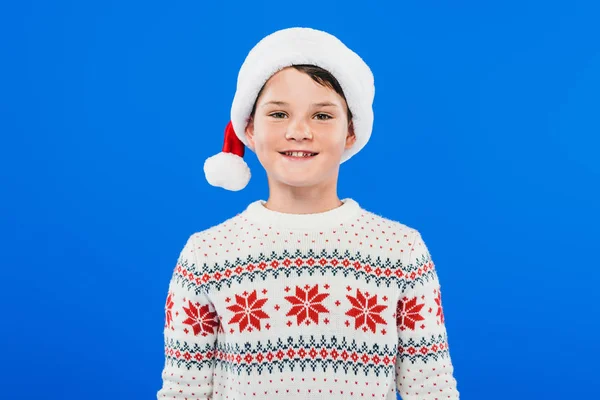 Vorderseite des lächelnden Kindes mit Weihnachtsmütze und Pullover isoliert auf blauem Grund — Stockfoto