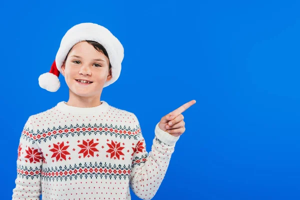 Vista frontal del niño sonriente en sombrero de santa señalando con el dedo aislado en azul - foto de stock