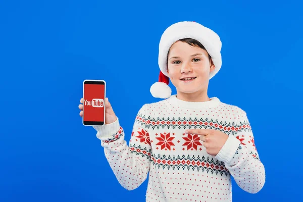 KYIV, UKRAINE - SEPTEMBER 9, 2019: front view of smiling kid in santa hat pointing with finger at smartphone with youtube app on screen isolated on blue — Stock Photo