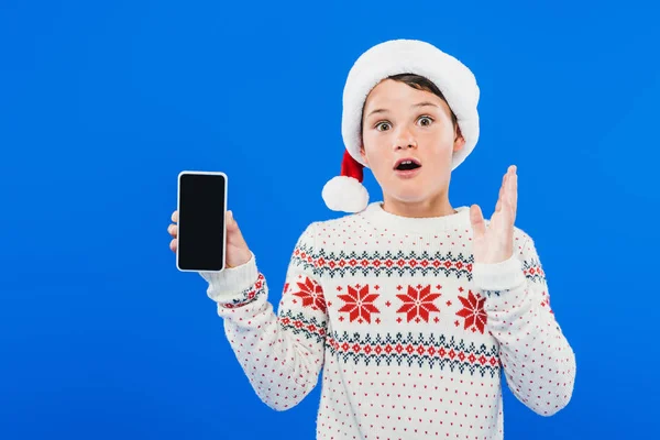 Front view of shocked kid in santa hat holding smartphone with blank screen isolated on blue — Stock Photo