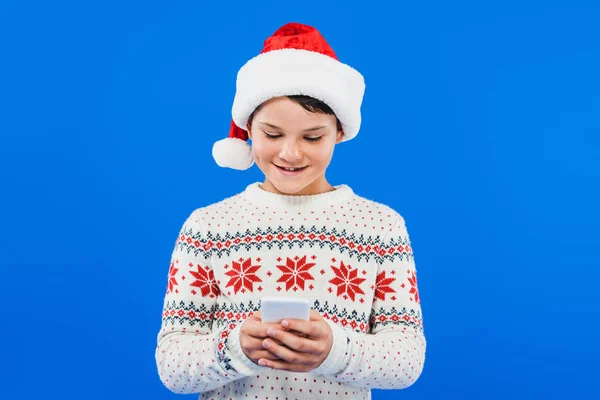 Frontansicht des Kindes mit Weihnachtsmütze und Smartphone isoliert auf blauem Grund — Stockfoto