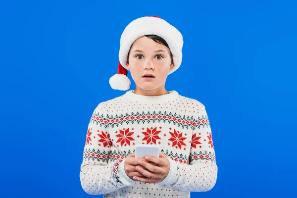 Frontansicht eines schockierten Kindes mit Weihnachtsmütze und Smartphone isoliert auf blauem Grund — Stockfoto