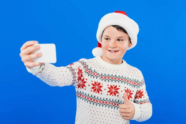 Sonriente niño en santa sombrero tomando selfie y mostrando el pulgar hacia arriba aislado en azul - foto de stock