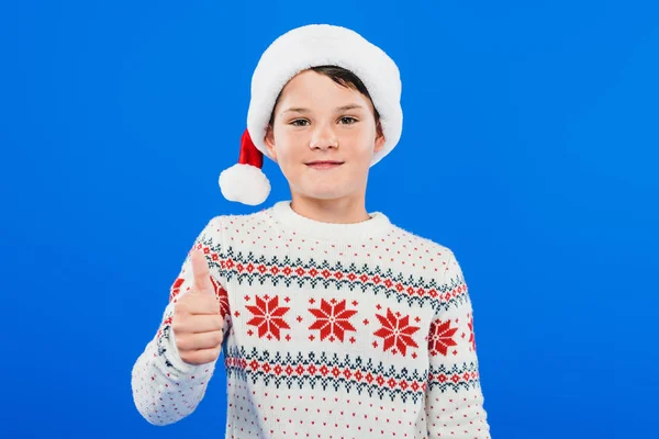 Vista frontale del bambino sorridente in cappello di Babbo Natale mostrando pollice in su isolato sul blu — Foto stock