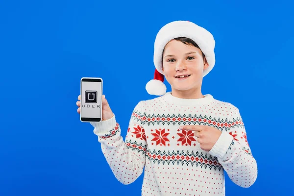 KYIV, UKRAINE - SEPTEMBER 9, 2019: front view of smiling kid in santa hat pointing with finger at smartphone with uber app on screen isolated on blue — Stock Photo