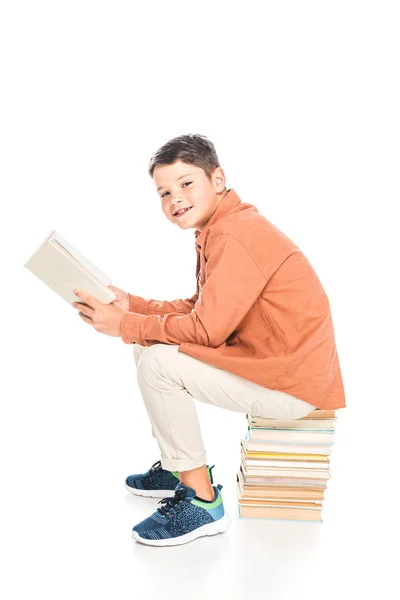 Bambino sorridente seduto sui libri e che legge sul bianco — Foto stock