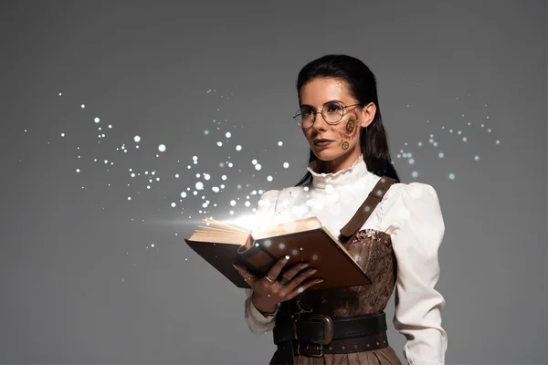 Mujer steampunk pensativo en gafas sosteniendo libro abierto con ilustración brillante hada aislado en gris - foto de stock