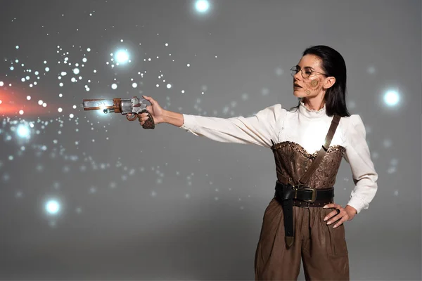 Steampunk woman shooting from revolver with hand on hip on grey background with glowing illustration — Stock Photo