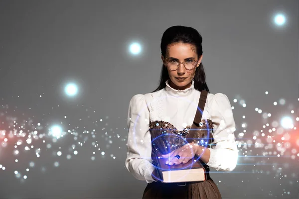 Pensive steampunk woman in glasses holding book with glowing illustration isolated on grey — Stock Photo