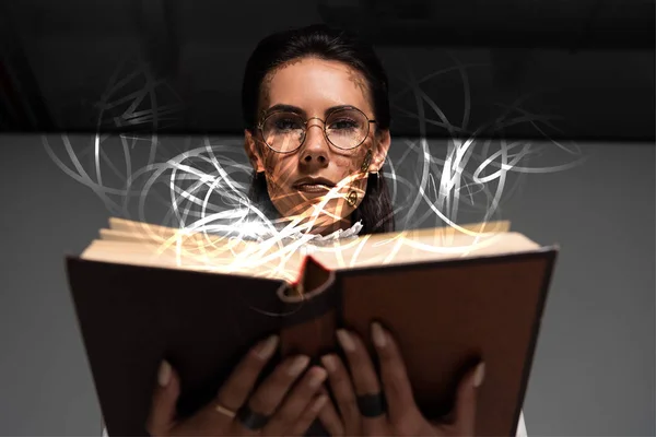 Low angle view of pensive steampunk woman in glasses reading book with glowing illustration above — Stock Photo