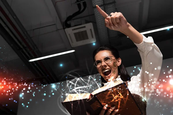 Low angle view of angry steampunk woman holding open book with fairy glowing illustration, screaming and pointing with finger — Stock Photo