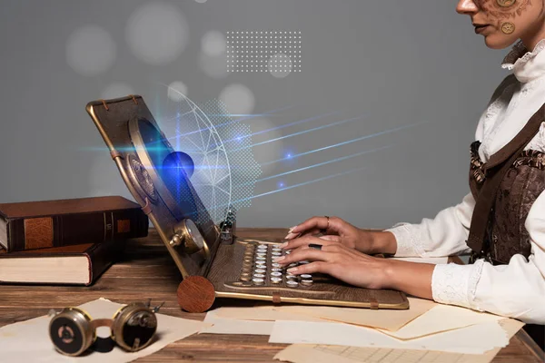 Cropped view of woman typing on steampunk laptop at table with glowing illustration isolated on grey — Stock Photo